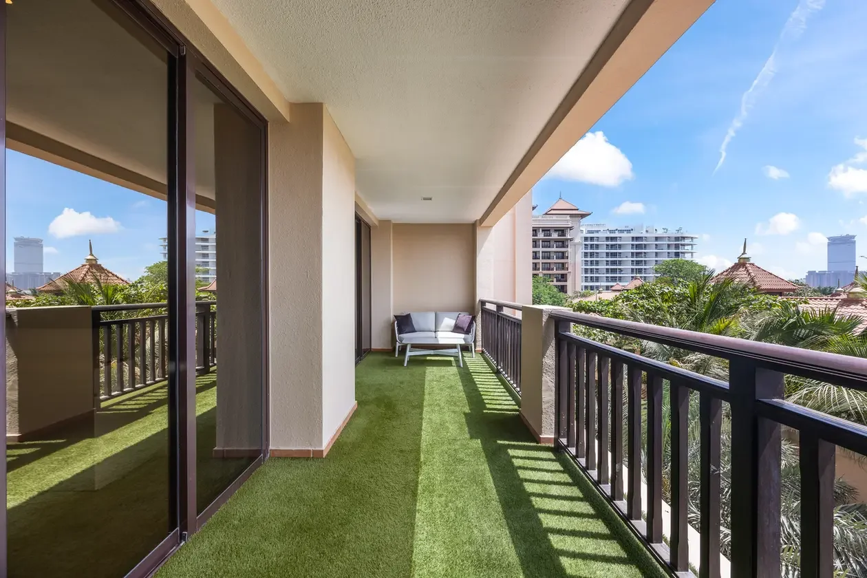 Balcony with artificial grass flooring, white outdoor sofa and purple cushions, and a city skyline in the distance.