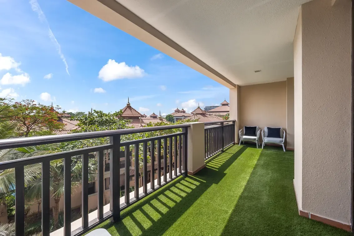 Balcony with green artificial turf, two white chairs with dark cushions, and a black railing overlooking red-tiled rooftops under a blue sky.