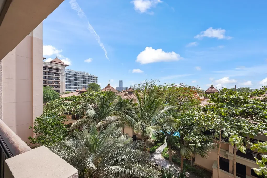 Balcony view of a tropical resort with palm trees, beige buildings, and a bright blue sky above terracotta rooftops.