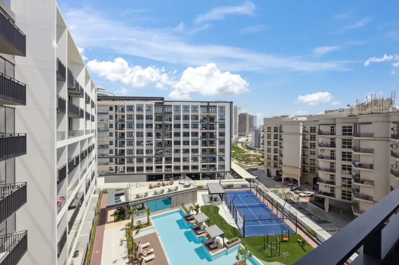 View from a balcony of a modern residential complex: white apartment buildings, a blue pool area with lounge chairs, and a fenced tennis court beneath a bright blue sky.