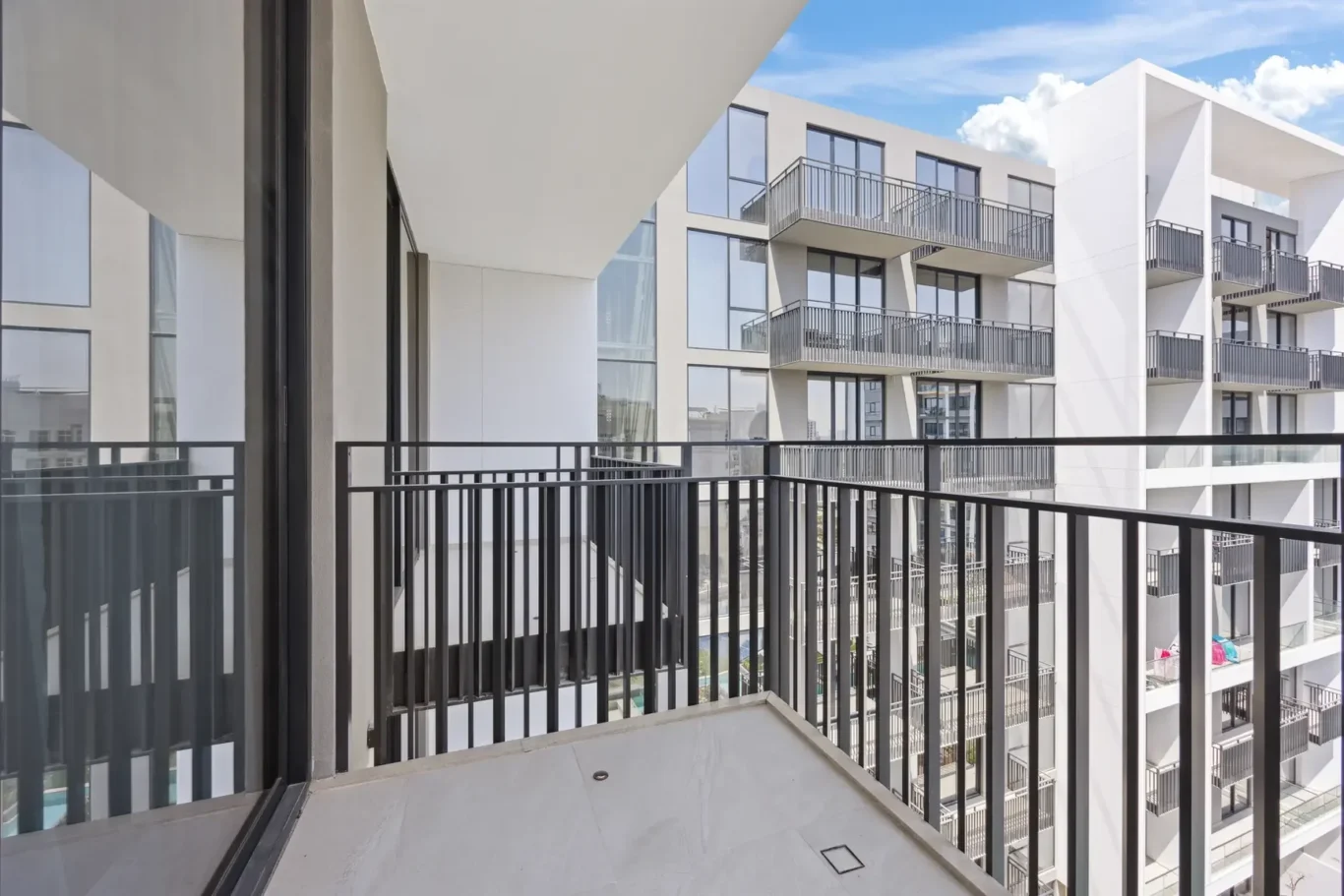 View from a modern apartment balcony: white building with glass windows and black railings, blue sky in the background.