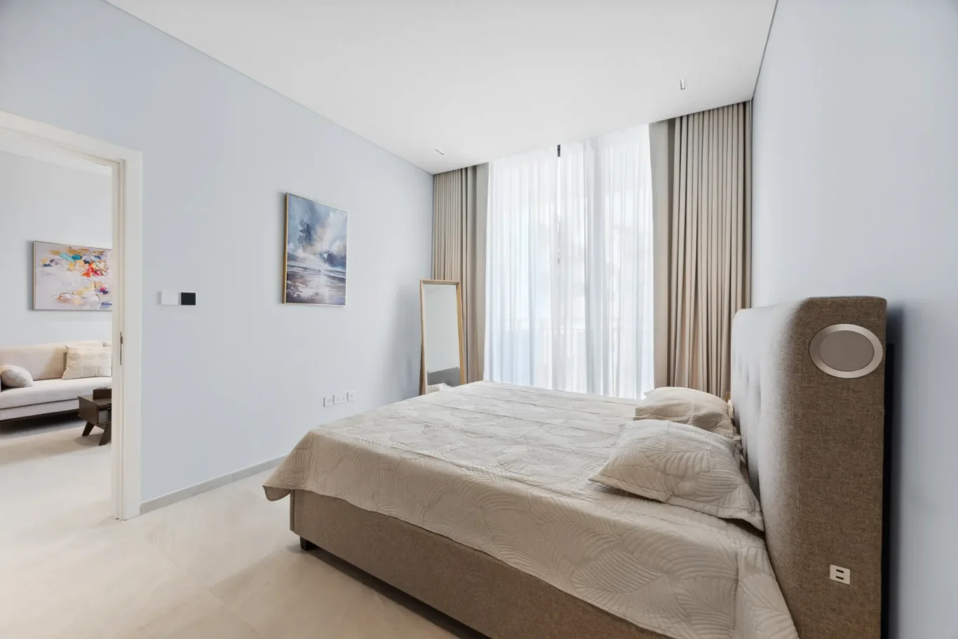 Bright modern bedroom with a large beige upholstered bed, light blue walls, and neutral bedding; artwork on the wall and a leaning mirror near the window.