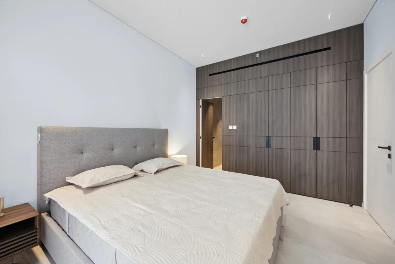 Modern bedroom with a grey upholstered bed, white quilted bedding, and a tall dark wood built-in wardrobe along the right wall