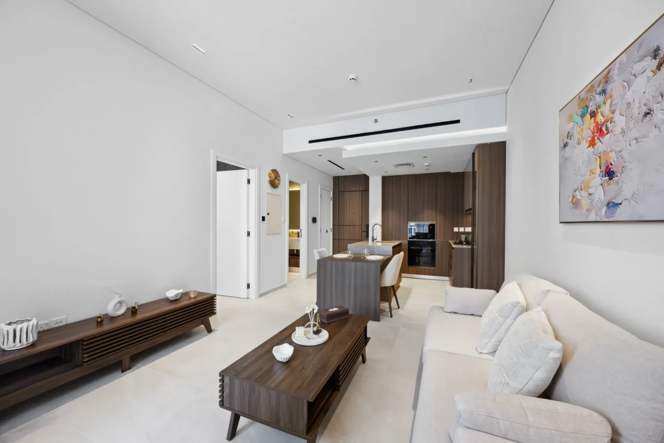 Open-concept living area with a beige sectional, two dark wood coffee tables, wall art, and a back kitchen with wooden cabinets.