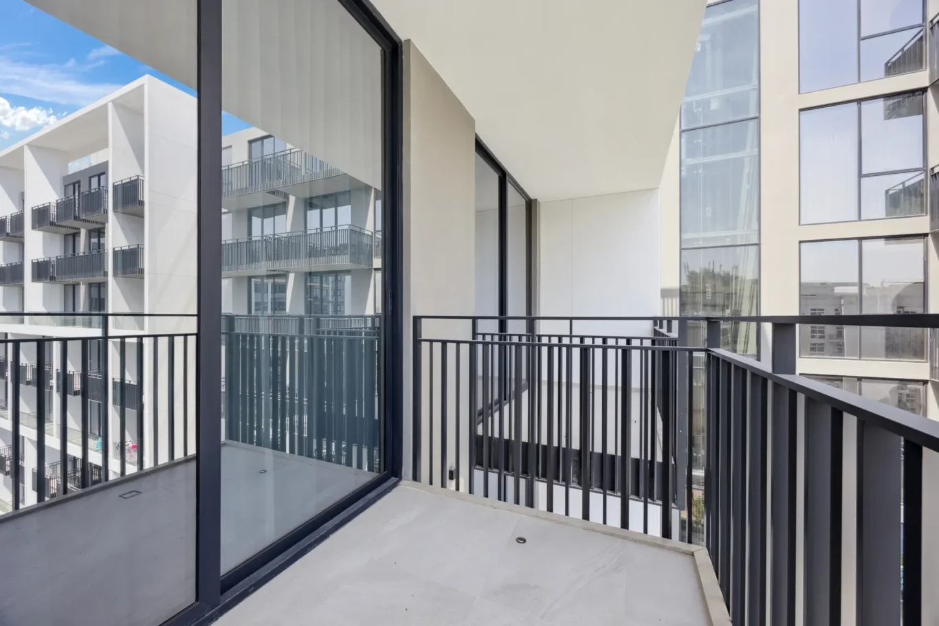 Balcony of a modern apartment building with glass walls and black metal railing, blue sky visible.