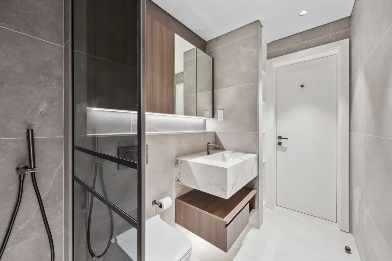 Modern gray-tiled bathroom with a glass shower enclosure, marble sink, and wooden vanity beneath a large mirror. The white door is at the far end.