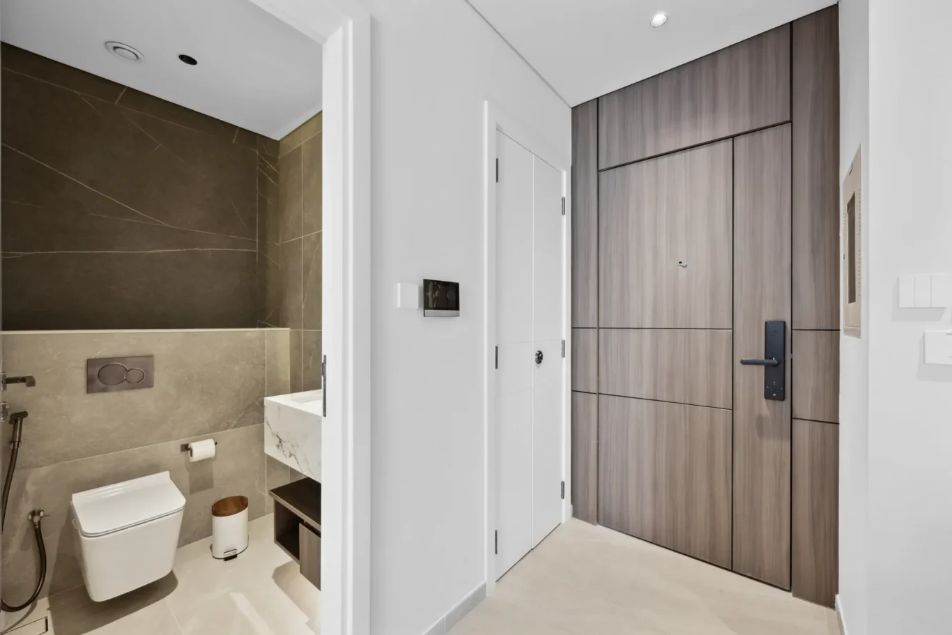Modern bathroom corner with wall-mounted toilet, grey marble walls, and a white vanity; doorway leads to a wood-panelled entry area.