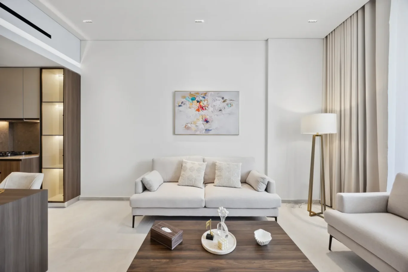 Modern living room with a light-gray sofa, abstract painting on the white wall, and a tall gold-floor lamp beside beige curtains; open kitchen area on the left.
