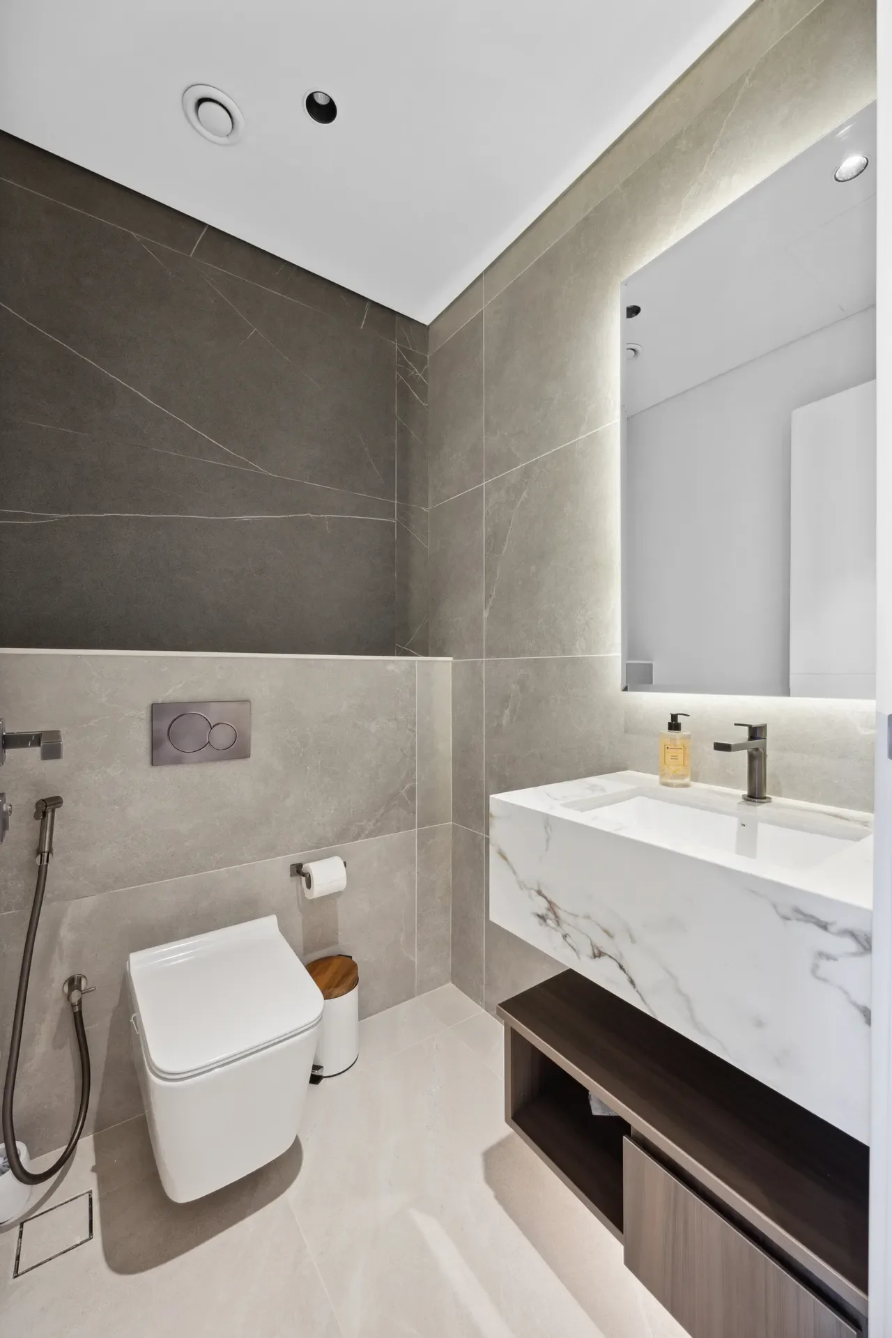Modern gray marble bathroom with wall-mounted toilet, bidet spray, and a marble vanity under a backlit mirror.