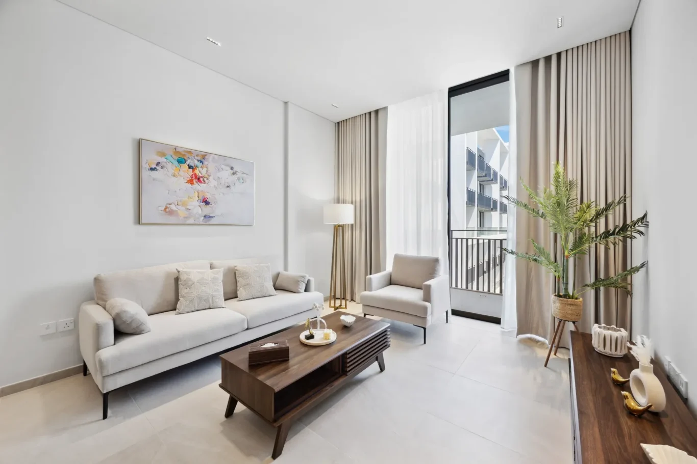 Modern living room with a cream sofa and armchair, wood coffee table, abstract wall art, and large glass doors with beige curtains.