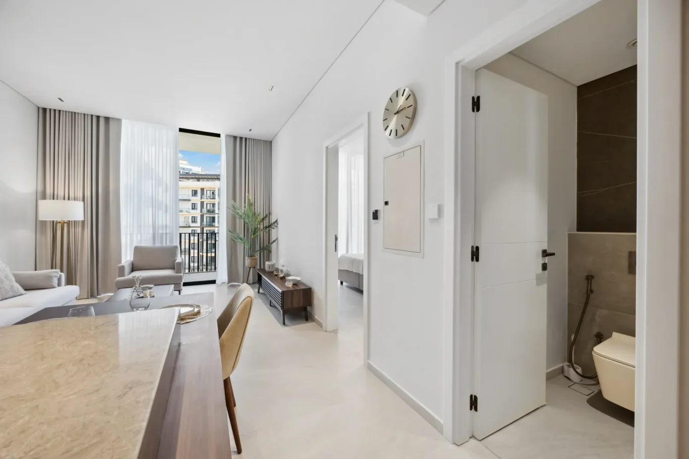 Bright living room with beige furniture and large windows with sheer curtains, overlooking a city building.