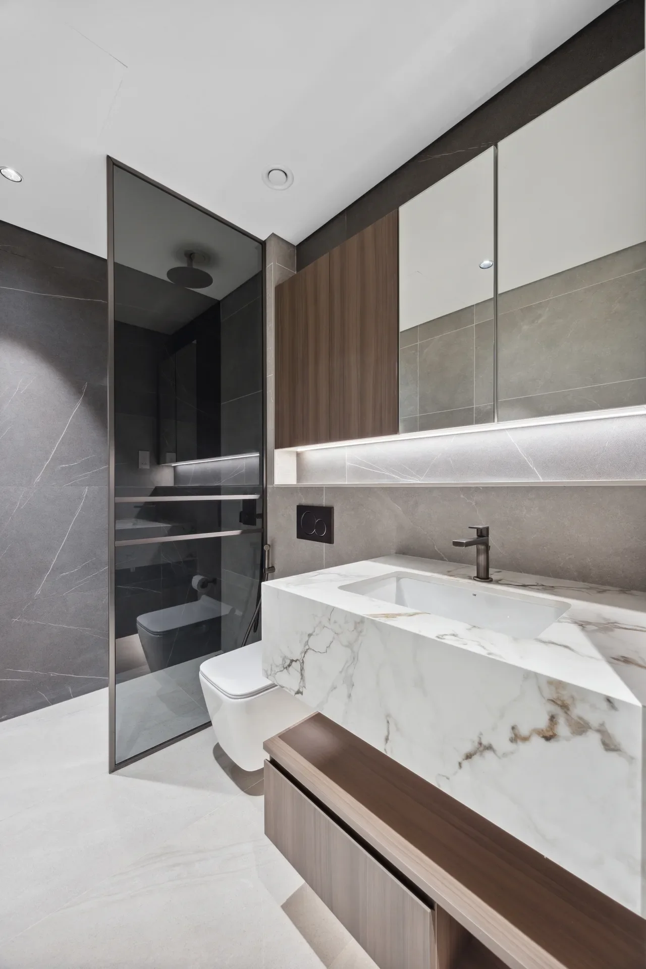 Modern bathroom with marble vanity, gray tile walls, and wooden cabinet panels; glass shower enclosure on left and wall-mounted faucet.