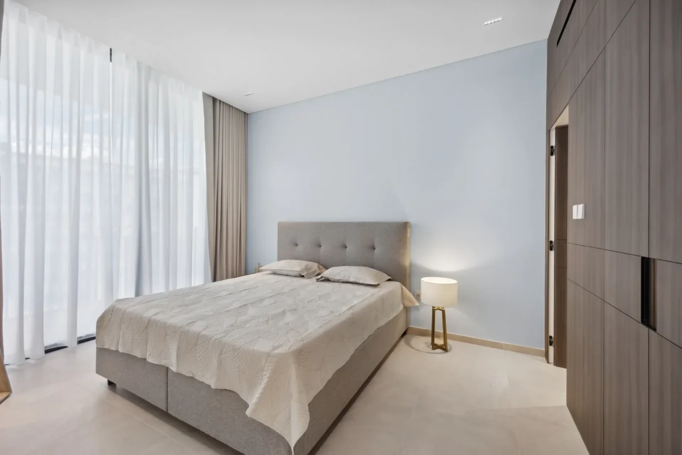 Modern bedroom with a gray upholstered bed, light blue walls, and a large wooden wardrobe on the right.