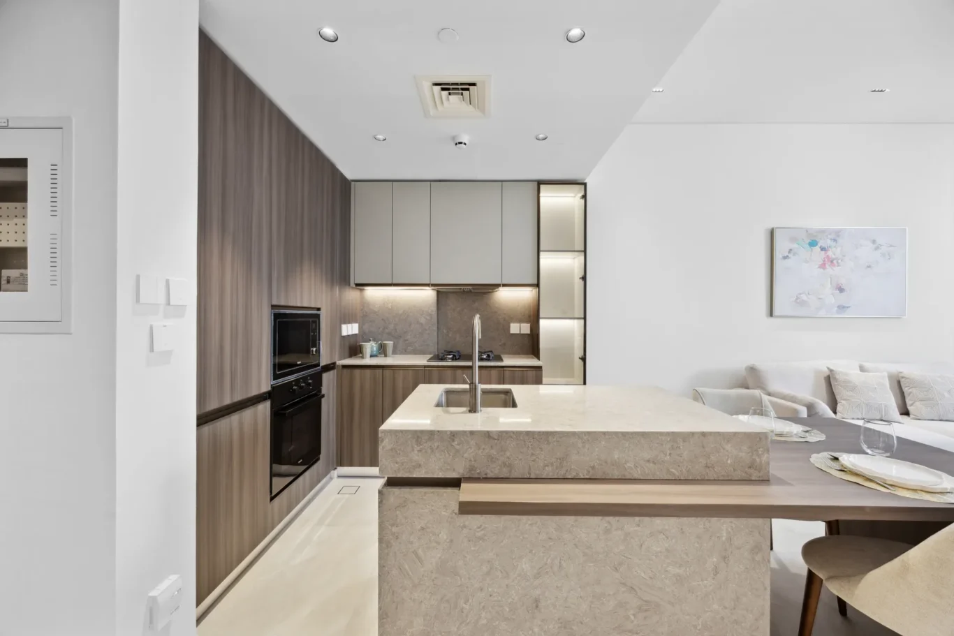 Modern kitchen with a large beige marble island, gray wall cabinets, and built-in appliances in an open living area.
