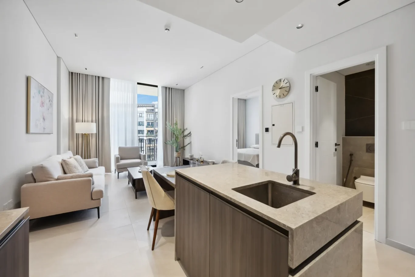 Open-plan living area with beige sofa, armchairs, coffee table, and a marble kitchen island in the foreground; large windows with sheer and gray curtains reveal a city view.