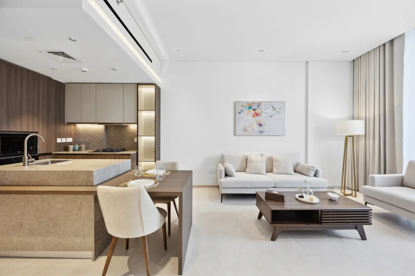 Open-plan living area with a beige kitchen island, light cabinets, and a dining table near a white sofa under an abstract painting.