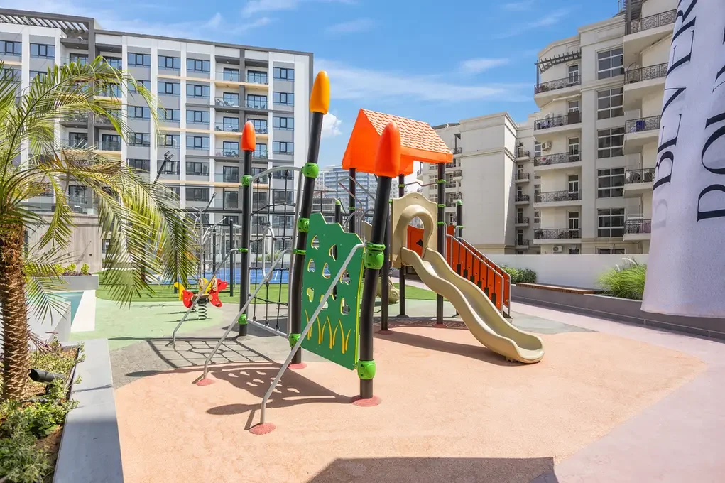 Colorful children's playground with slides and climbing wall in a courtyard surrounded by modern apartment buildings on a sunny day.