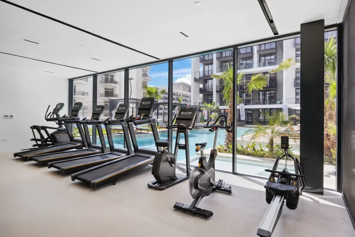 Modern hotel gym with floor-to-ceiling windows, row of treadmills, and stationary bikes facing an outdoor pool area with palm trees.