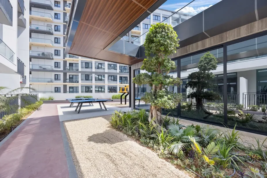 Outdoor courtyard of a modern apartment complex with a ping-pong table, gravel path, and lush plants.