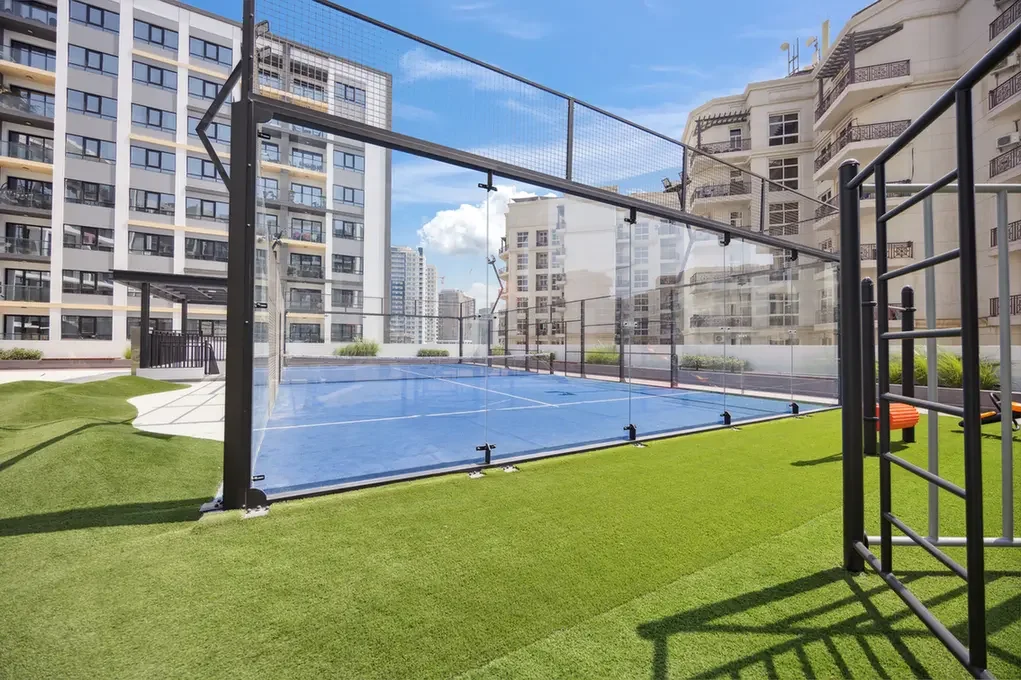 Rooftop outdoor sports court with a blue surface enclosed by glass panels, surrounded by green artificial turf and modern apartment buildings on a sunny day.