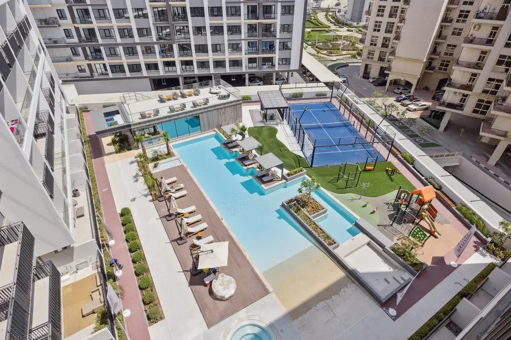Aerial view of a modern apartment complex courtyard with a large pool, sun loungers, shaded seating, a kiddie pool, and a playground area nearby, surrounded by tall buildings.