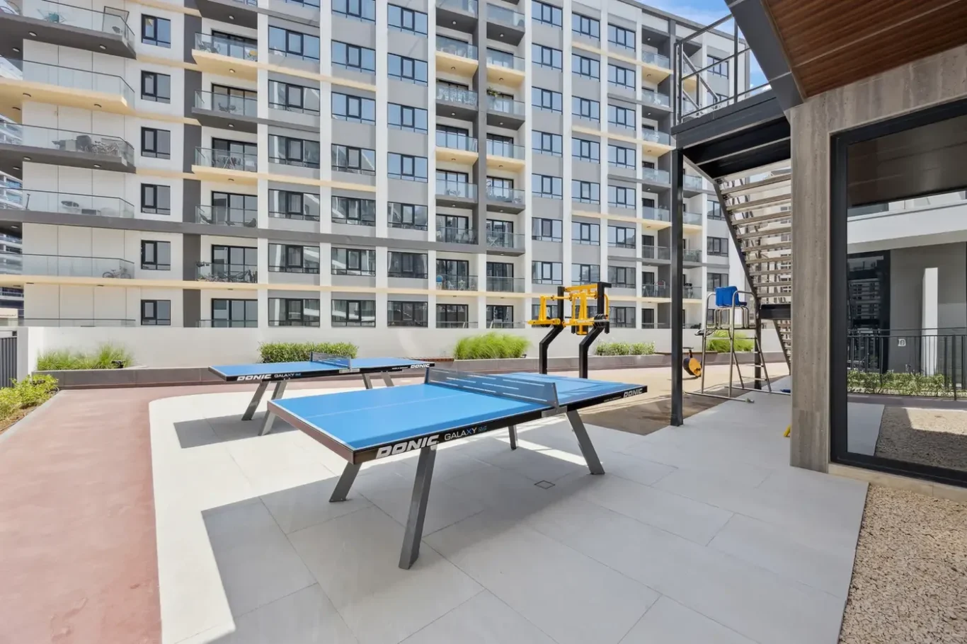 Outdoor recreation area with two blue Donic Galaxy ping-pong tables and a yellow workout frame in front of a modern apartment building with glass balconies.