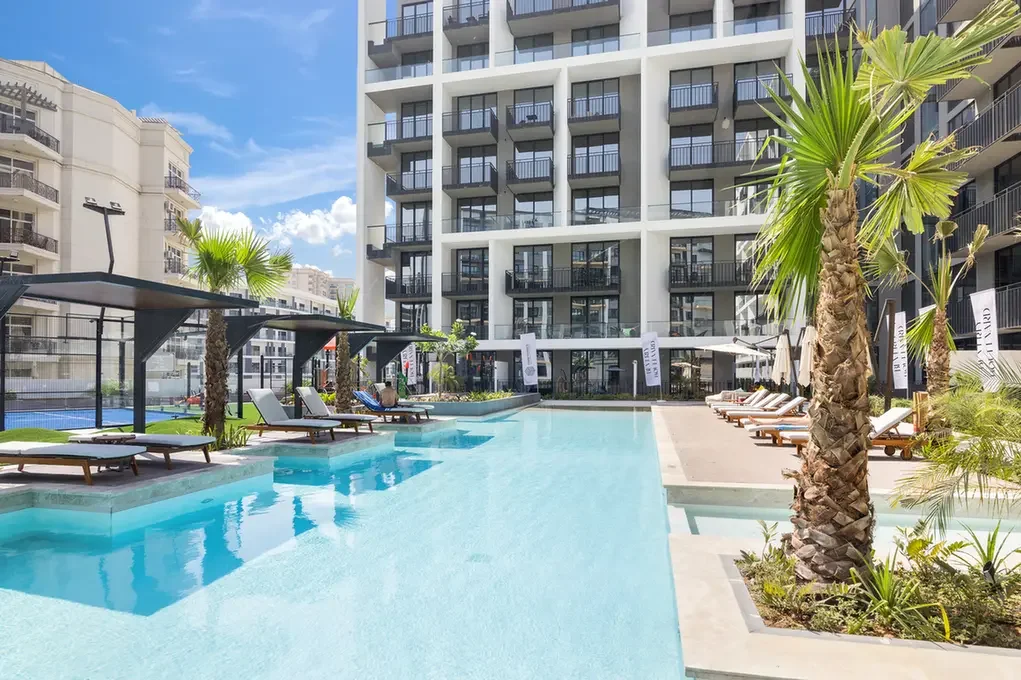 Poolside area at a modern hotel with blue water, palm trees, and a white multi-story building with balconies in the background.