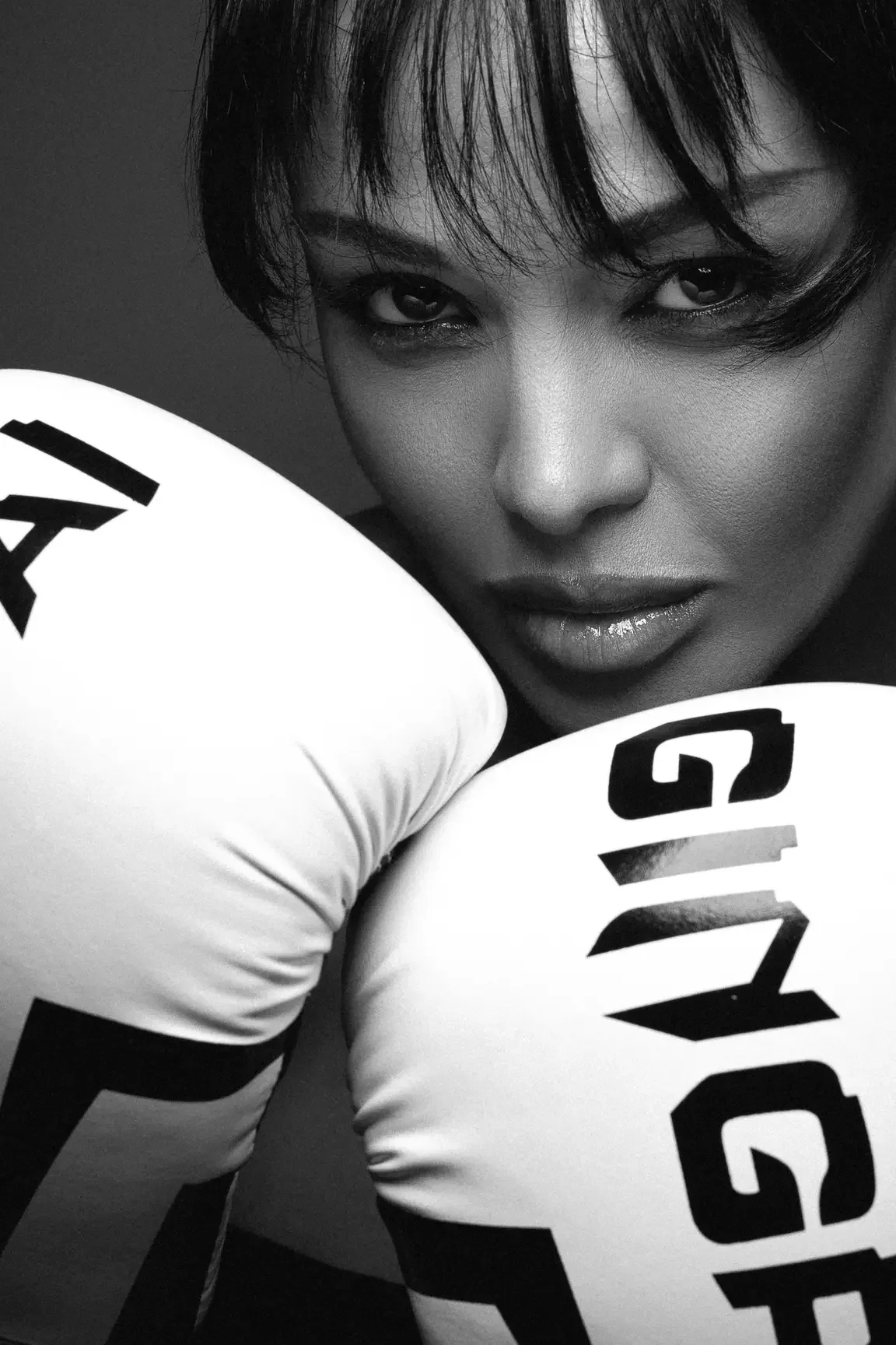 Close-up of a woman with boxing gloves near her face, looking at the camera in a black-and-white photo.
