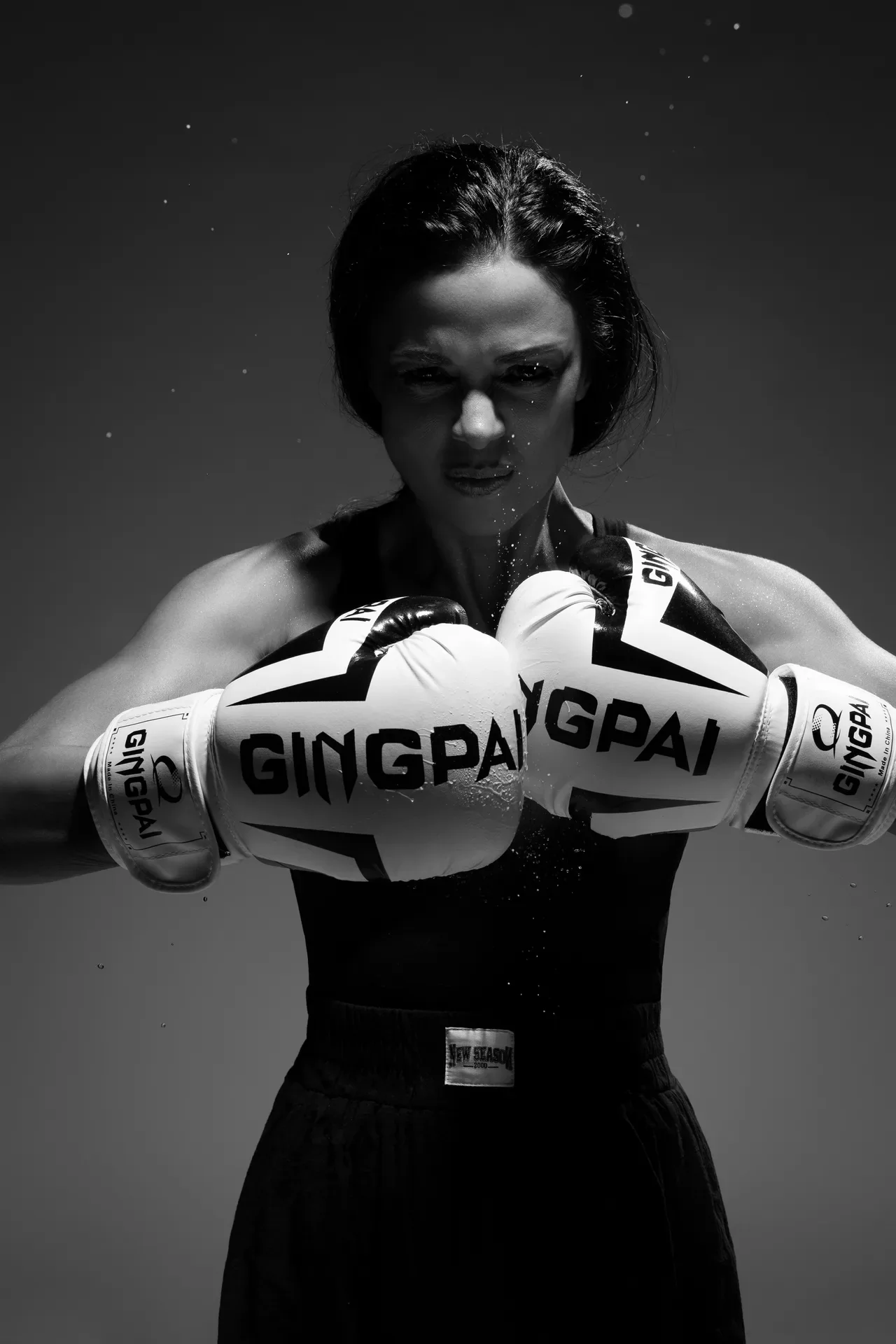 Female boxer in a fighting stance with white gloves labeled GING GAI, black-and-white photo.