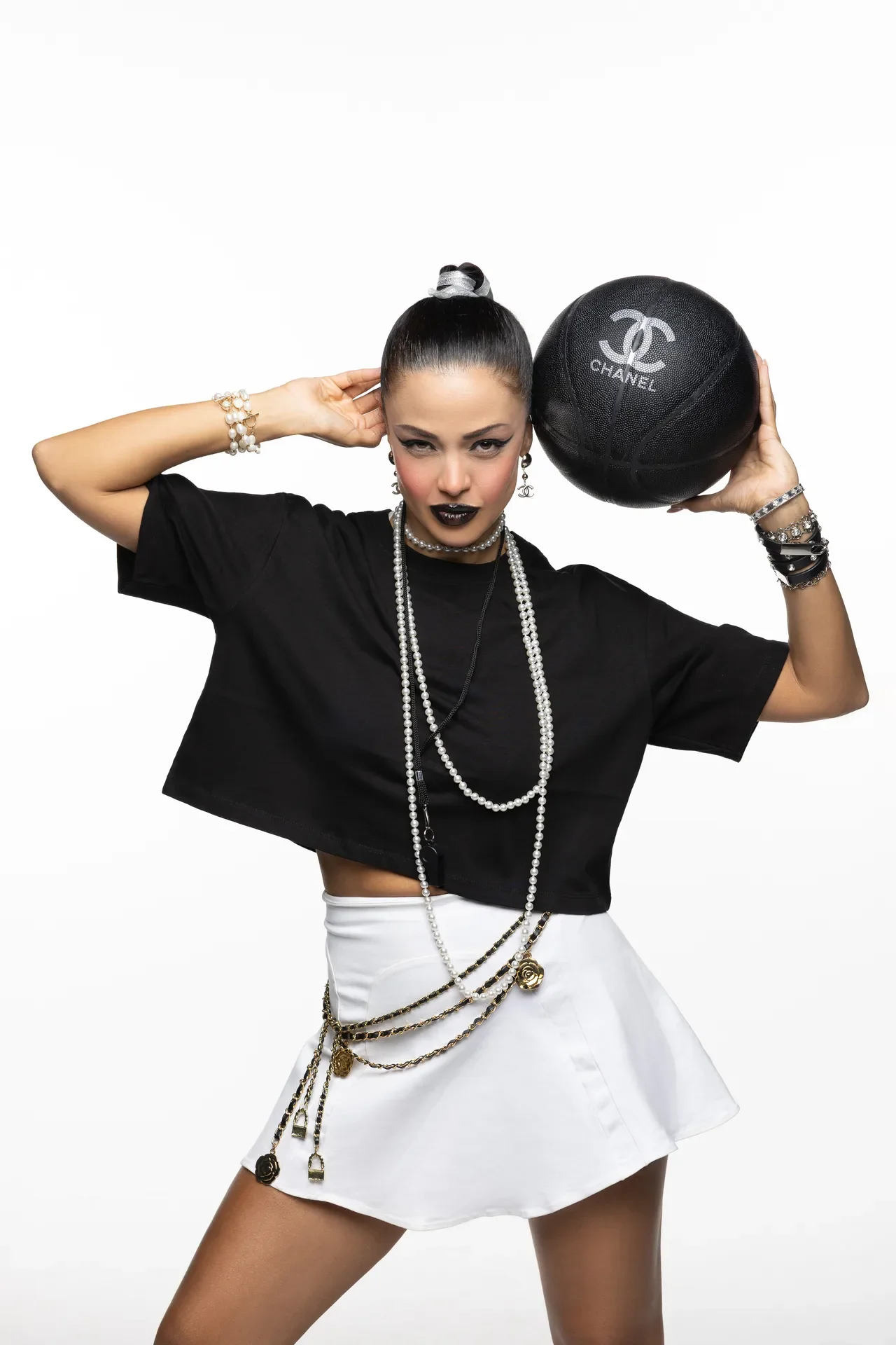 Fashion model posing with a black Chanel basketball, wearing a black crop top, white skirt, and multiple pearl necklaces.