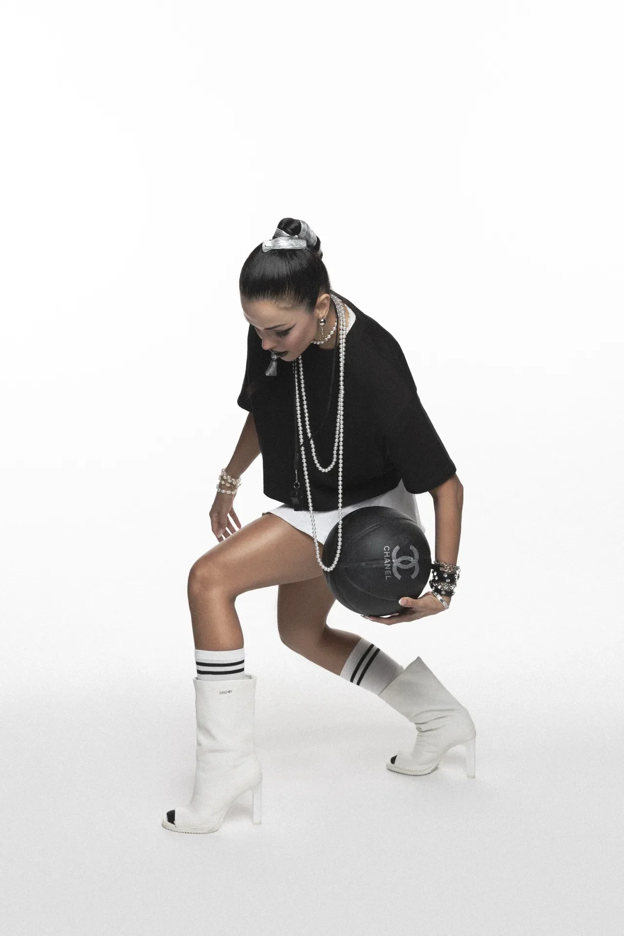 Woman wearing a black top, multiple pearl necklaces, and white knee-high boots bends forward while holding a round black Chanel handbag with the brand logo visible.