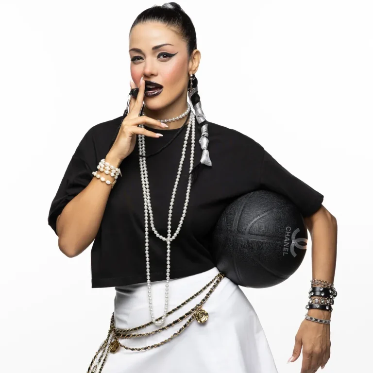 Woman with black top and white skirt, holding a basketball under her arm, finger to lips and bold pearl accessories.
