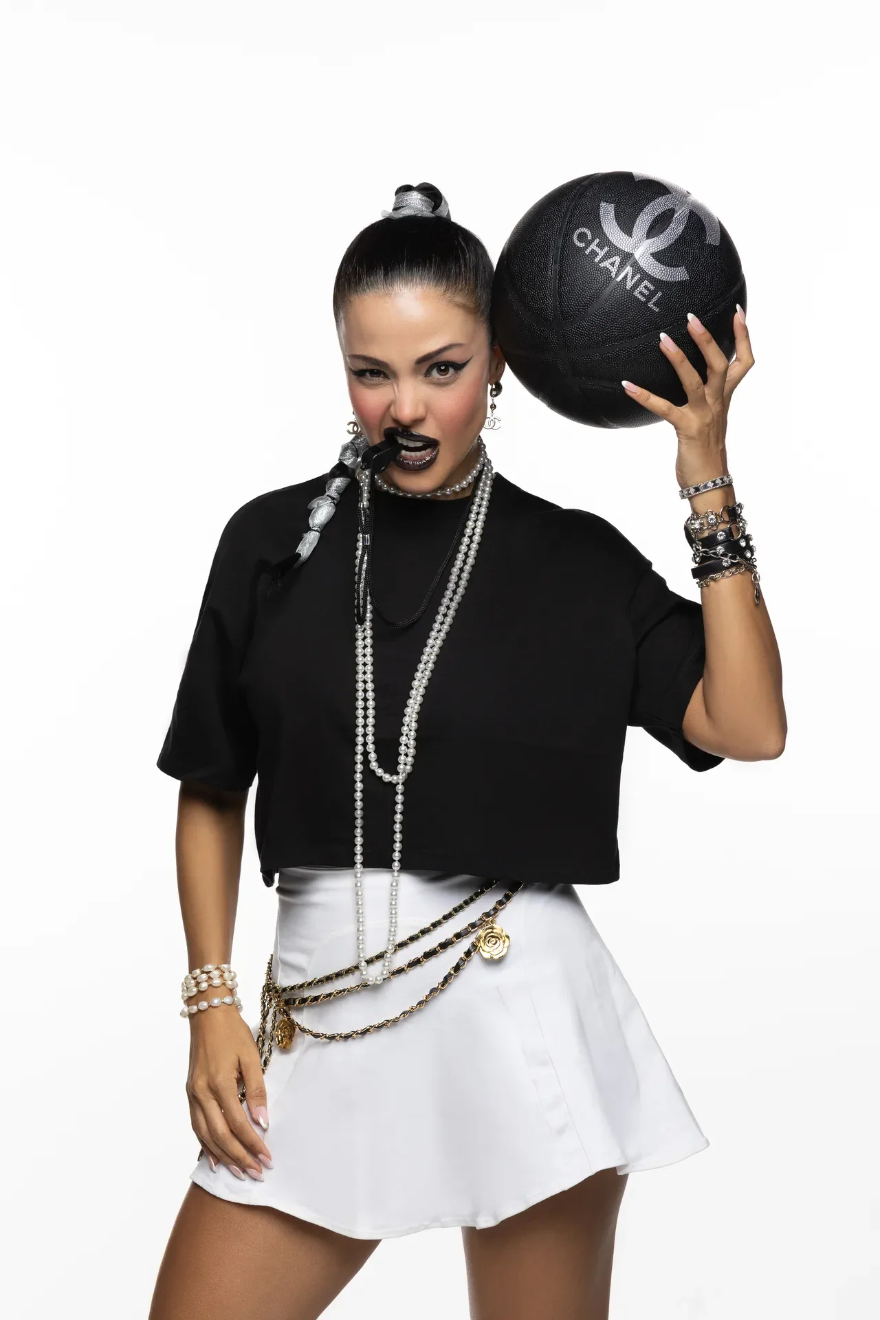 Stylish woman in a black top and white skirt, wearing layered pearls, holds a black Chanel basketball beside her head.
