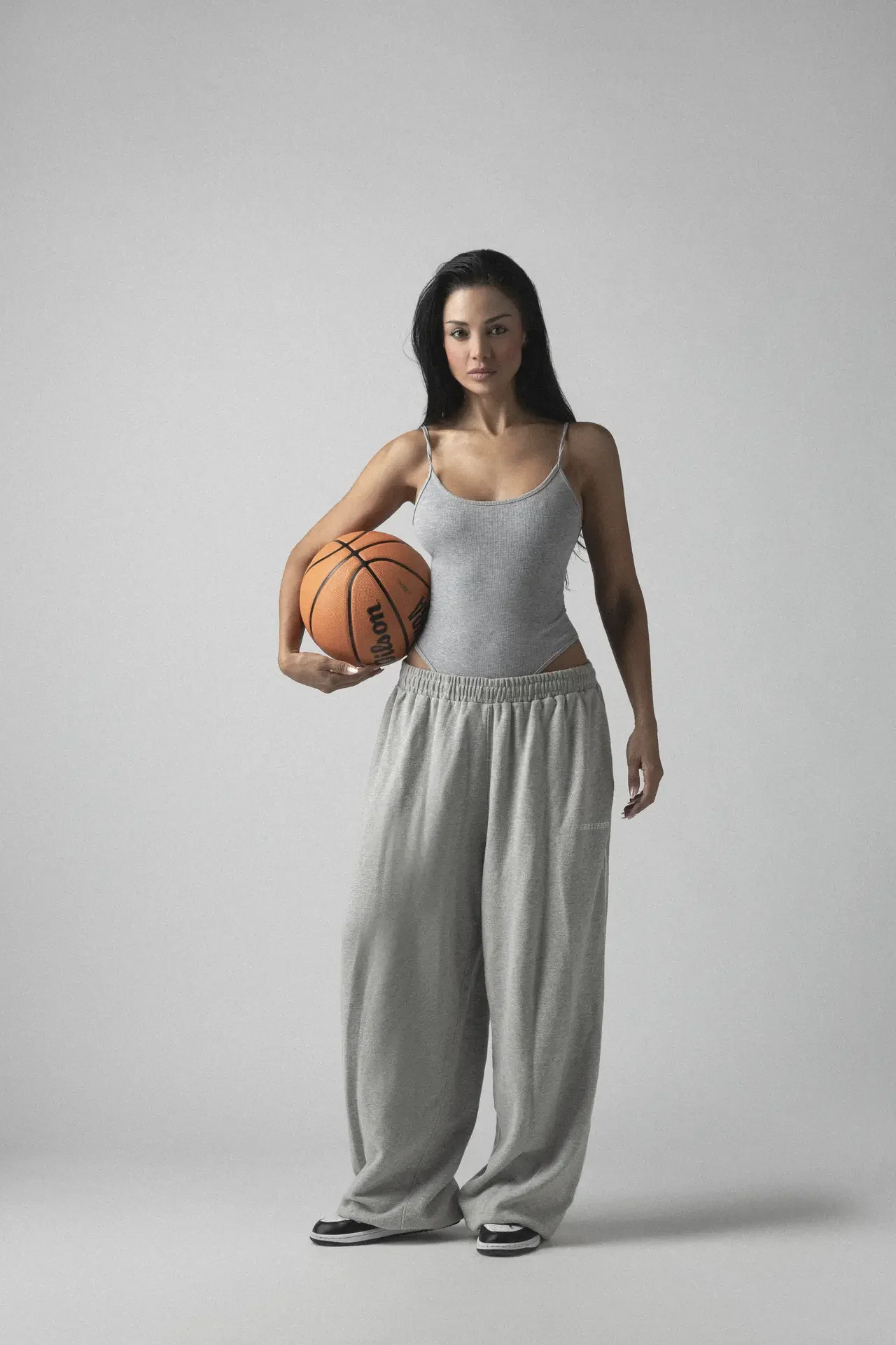 Athletic woman in a gray tank top and baggy sweatpants stands against a gray studio backdrop, holding a basketball under her left arm.