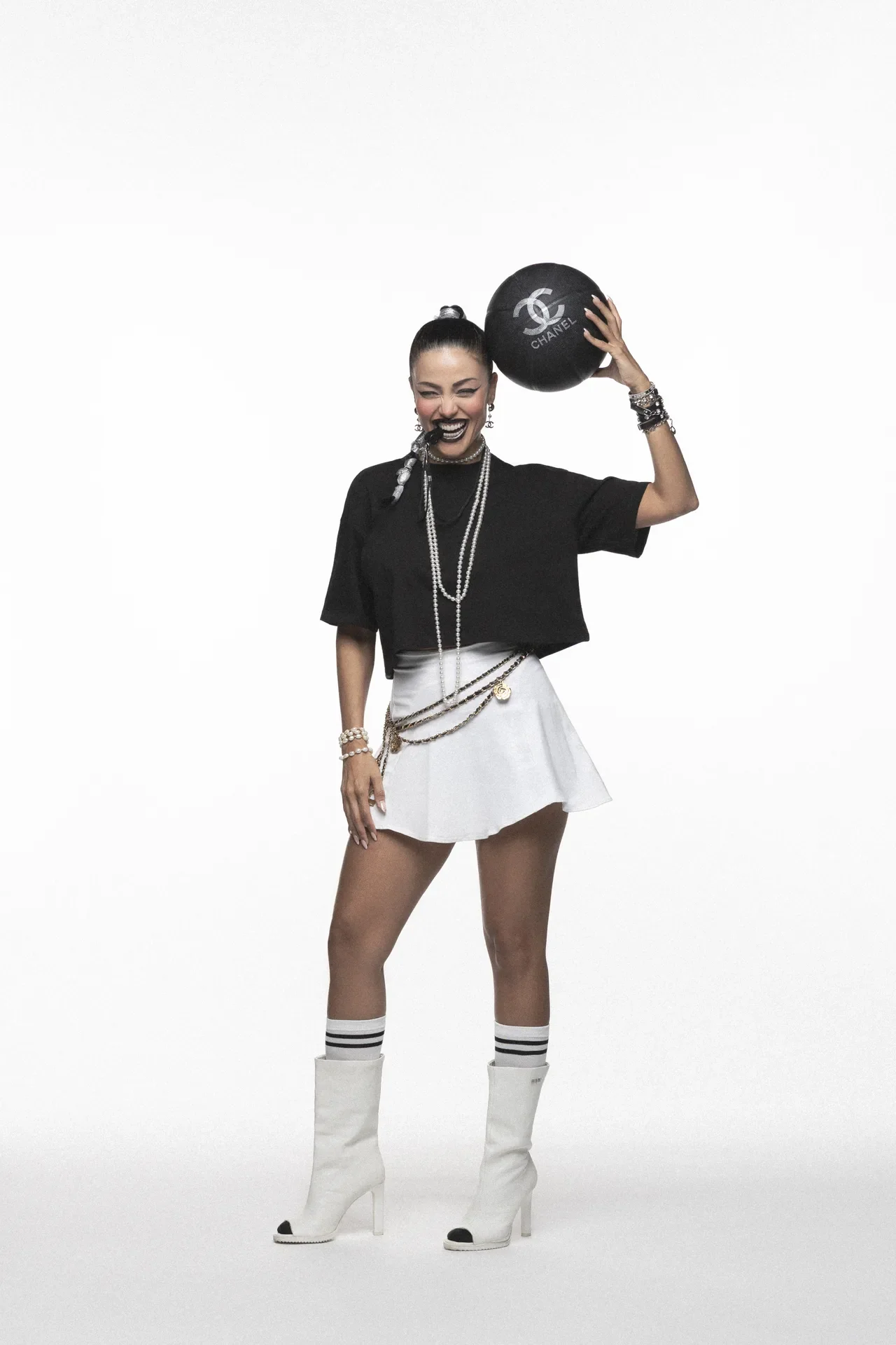 Model in a black cropped top, white mini skirt, white knee-high boots, and layered pearls, holding a black Chanel ball overhead with a smile.",
