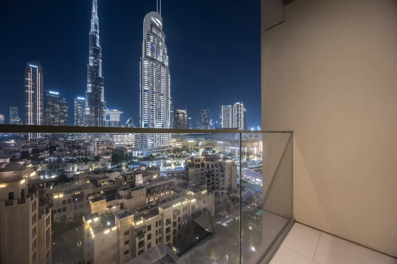 Nighttime city skyline viewed from a balcony, with a glass railing in the foreground and tall illuminated skyscrapers across the horizon.