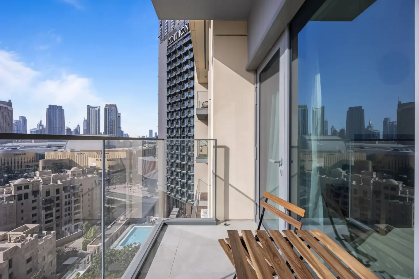 Balcony with glass railing overlooking a city skyline; a wooden folding chair on the tiled floor, with a pool visible below.