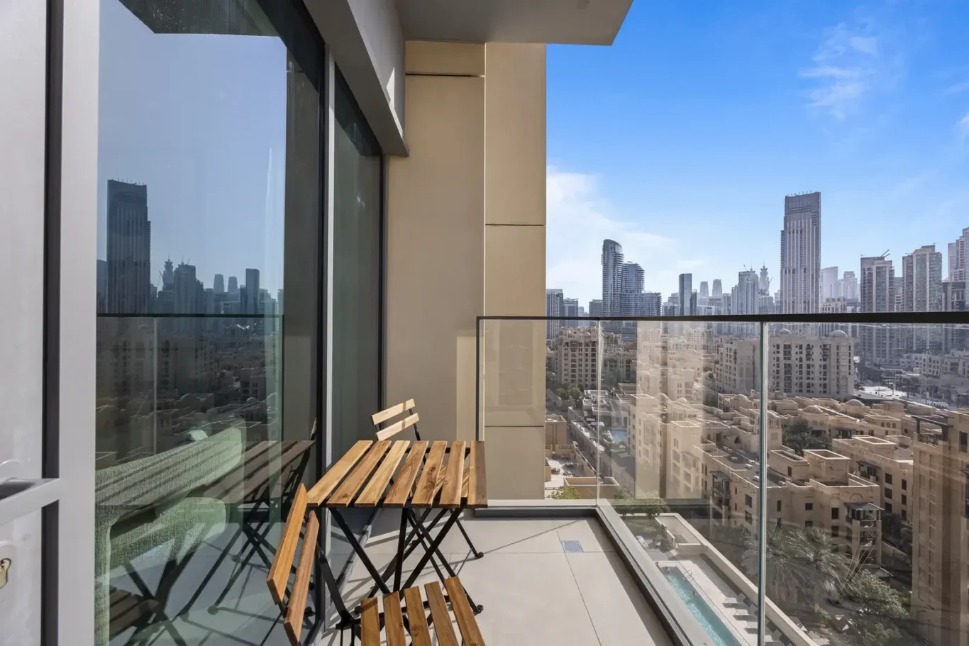 Balcony with a wooden table and chairs overlooking a city skyline of tall beige and glass buildings at noon.