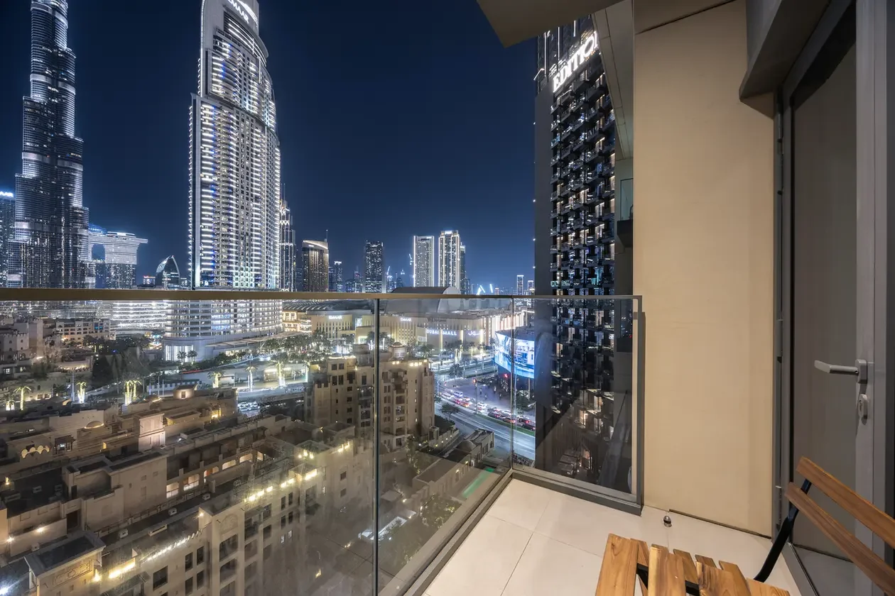 Nighttime cityscape view from a high-rise balcony with glass railing and illuminated skyscrapers.