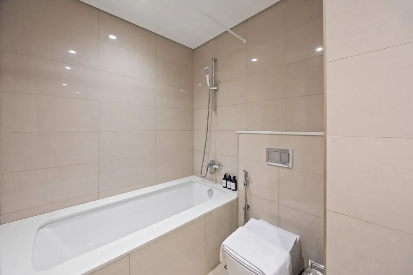 Modern beige-tiled bathroom with built-in bathtub, wall-mounted shower, and a towel on the toilet edge.