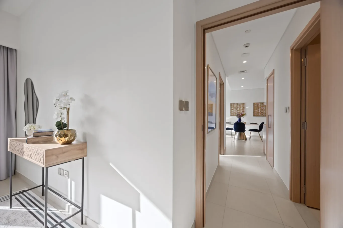 Hallway view into a dining area with a round table and blue chairs, seen through an open wooden doorway from a console table with books and a white orchid in the foreground