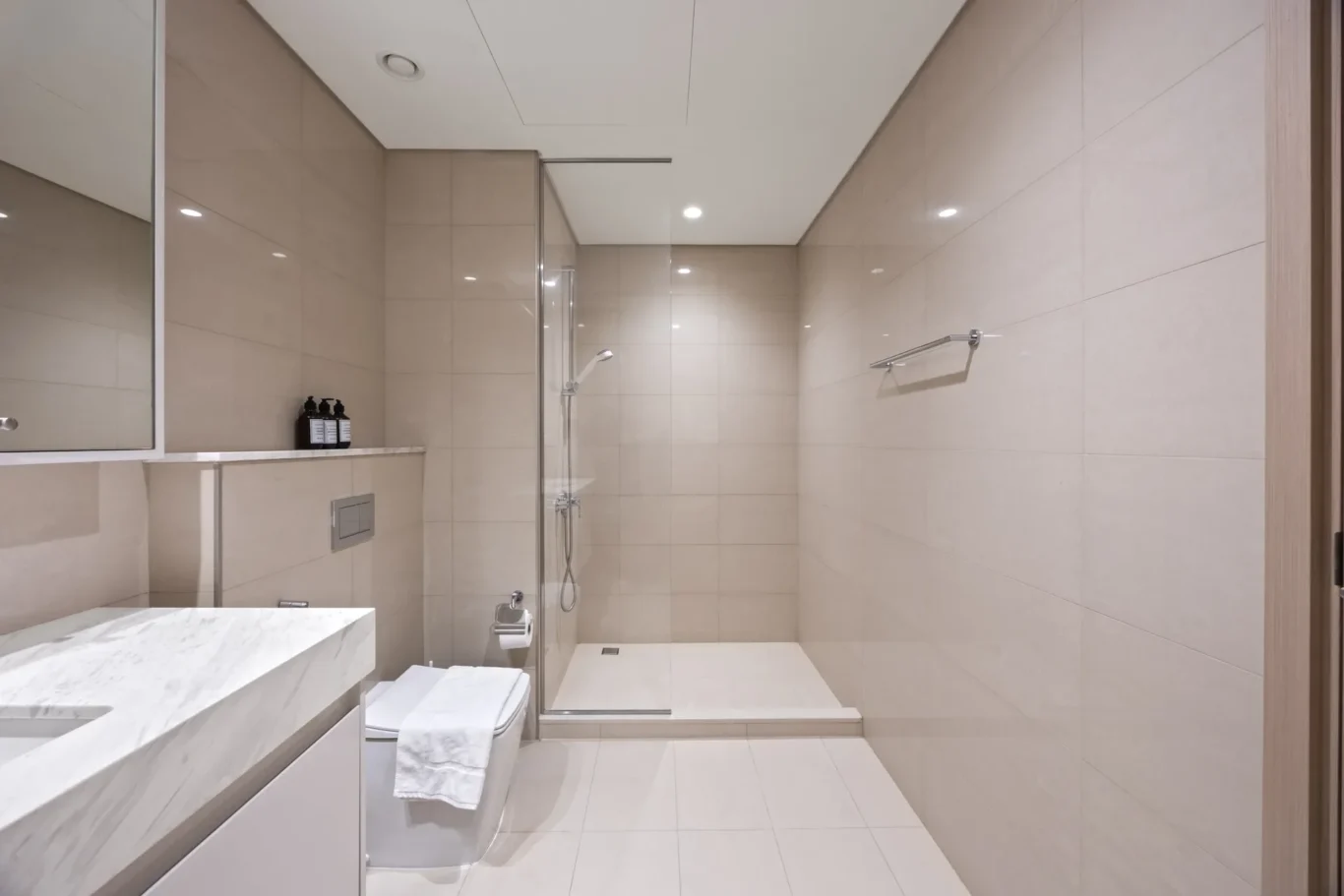 Modern beige-tiled bathroom with glass shower enclosure, marble vanity, and neatly folded towels