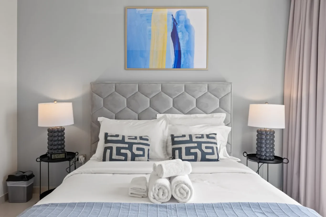 Hotel-style bedroom with tufted gray headboard, white bedding, abstract blue and yellow painting, and two glass lamps on each side.