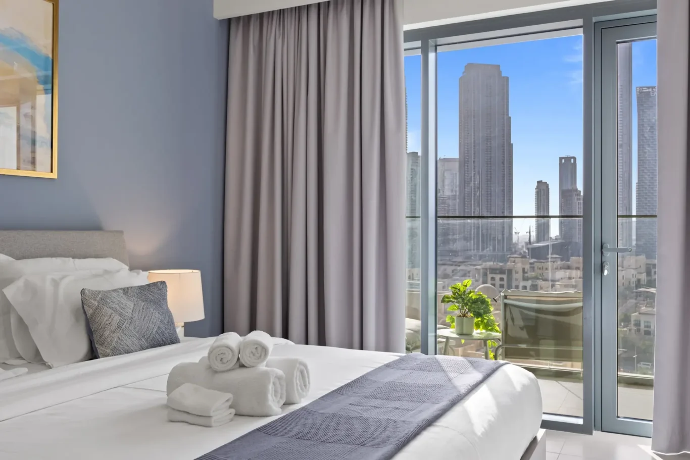 Hotel room with a neatly made bed, gray throw pillows, and rolled towels on the duvet; a balcony door reveals a city skyline.