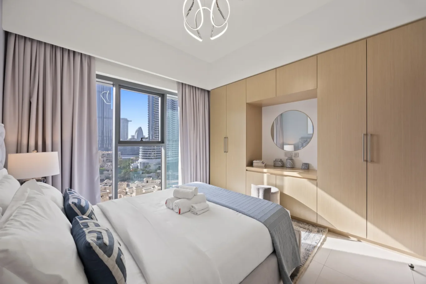 Hotel bedroom with white linens, blue patterned pillows, and folded towels on the bed; city skyline visible through large windows.