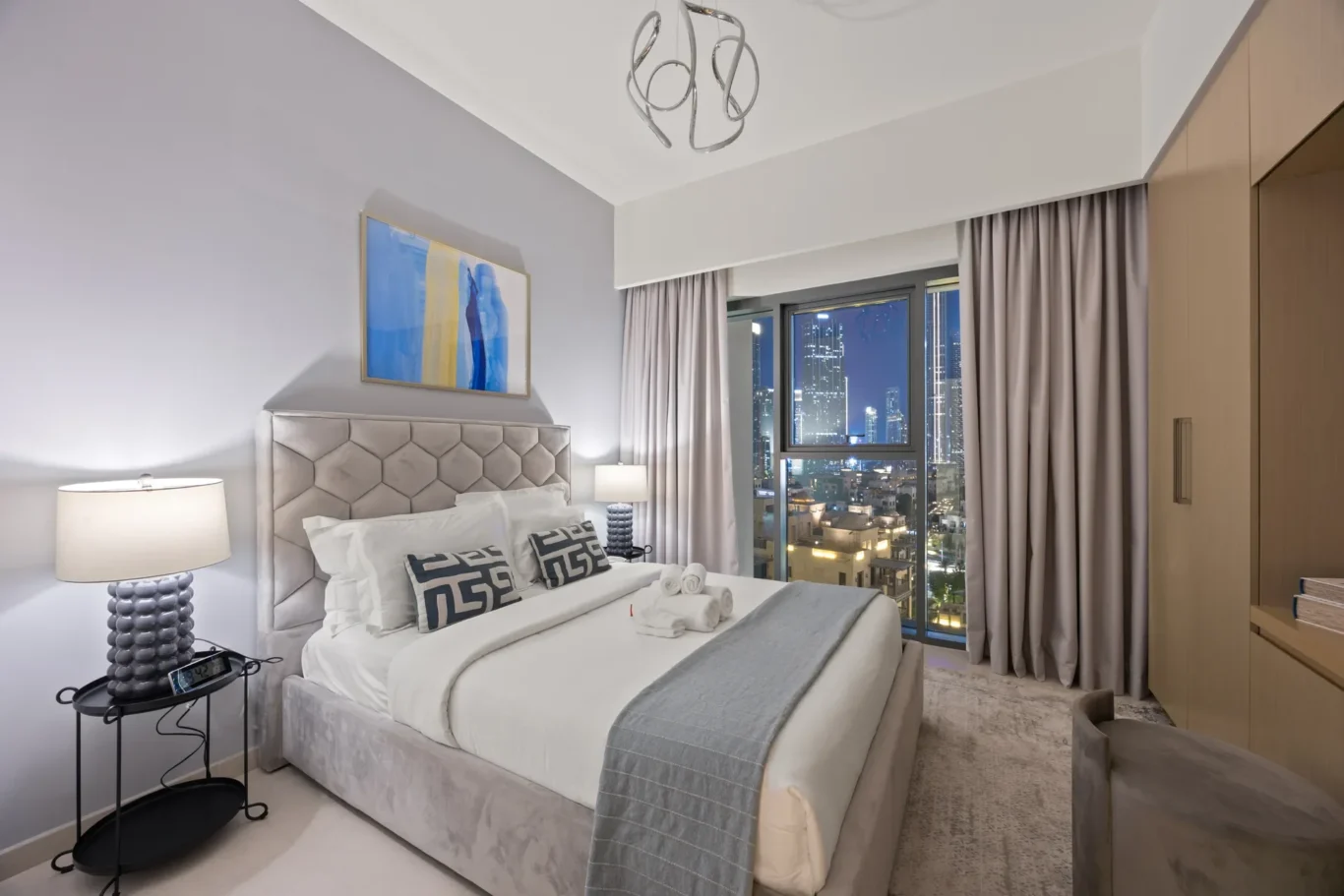 Hotel bedroom with a tufted gray headboard, white bedding, and a city skyline visible through a large window with gray curtains.