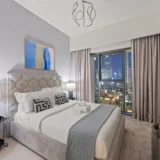 Hotel bedroom with a tufted gray headboard, white bedding, and a city skyline visible through a large window with gray curtains.