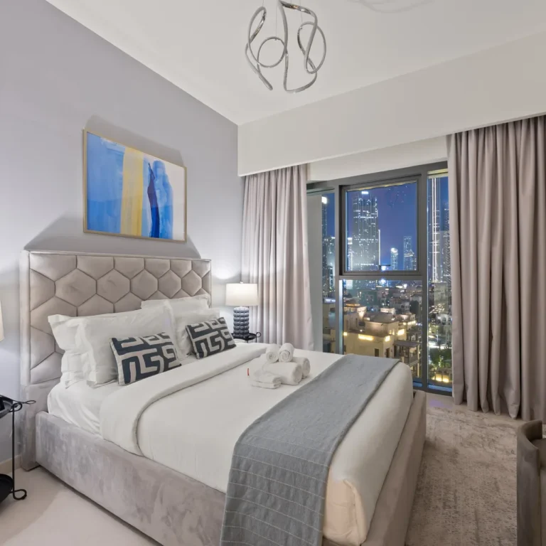 Hotel bedroom with a tufted gray headboard, white bedding, and a city skyline visible through a large window with gray curtains.