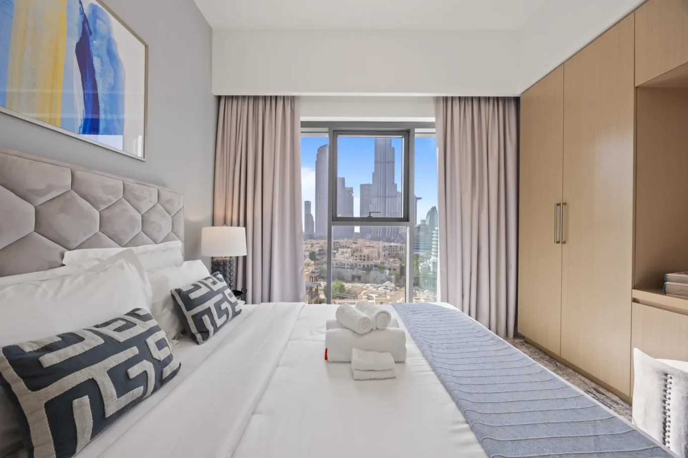 Modern hotel bedroom with a tufted white headboard, geometric-pattern pillows, and city skyline visible through a large window.
