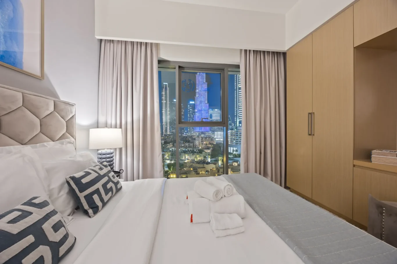 Hotel bedroom at night with a large window showing a lit city skyline; bed with a tufted beige headboard and white linens, decorative pillows, and a nightstand lamp on the left.